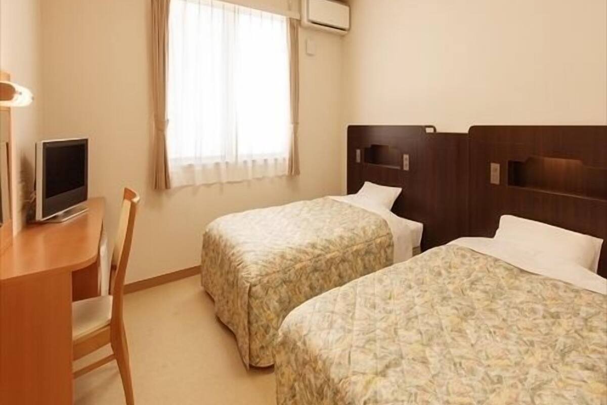 Ein Hotelzimmer mit zwei Betten, einem Schreibtisch mit Computer, einem Fenster mit Vorhängen und einer Deckenleuchte.