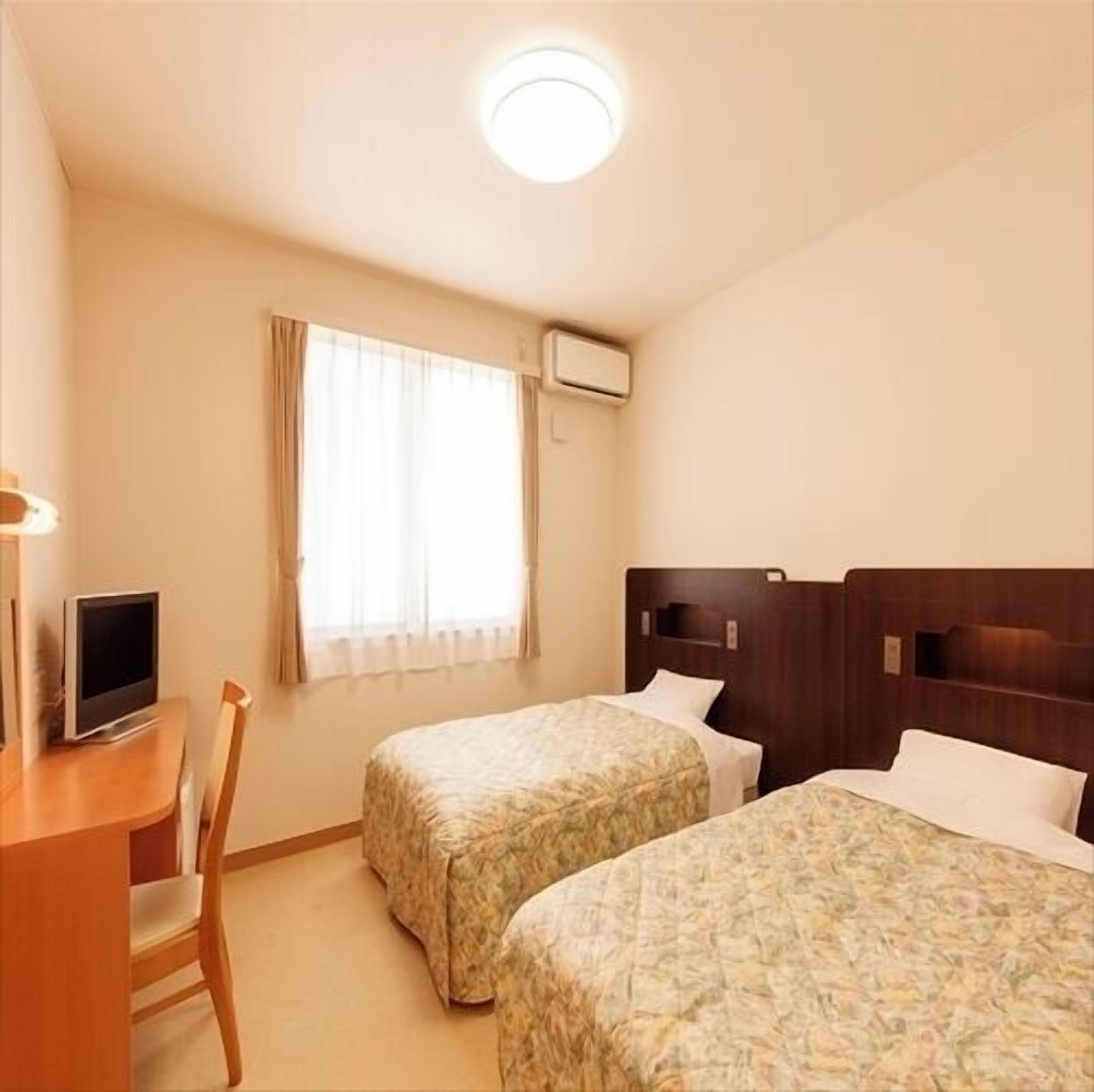 A hotel room with two beds, a desk with a television, a window with curtains, and an air conditioning unit.