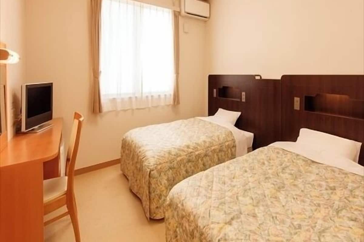 Ein Hotelzimmer mit zwei Betten, einem Schreibtisch mit Fernseher, einem Fenster mit Vorhängen und einer Klimaanlage.