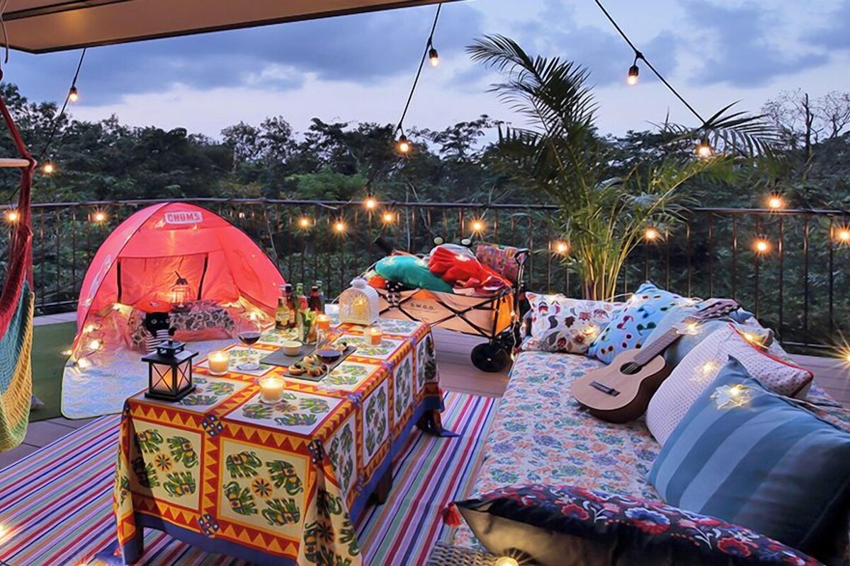 An outdoor seating area with a hammock, a red tent labeled 'CHUMS', a table with food and drinks, and a guitar.