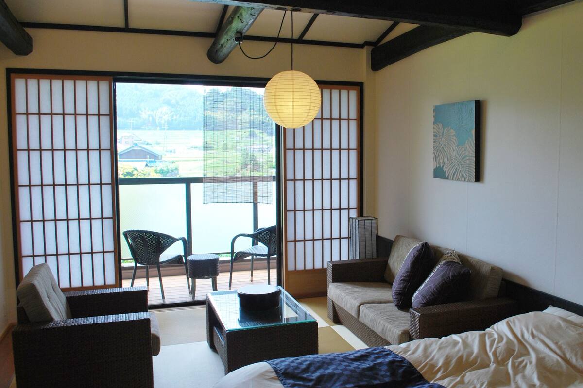 Une pièce de style japonais traditionnel, avec un sol recouvert de tatamis, une table basse, un canapé et une porte coulissante donnant sur un balcon.