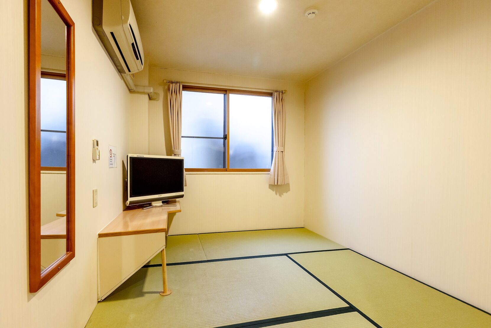 A room with tatami flooring, a television on a wooden stand, a window with curtains, and a ceiling-mounted air conditioner.