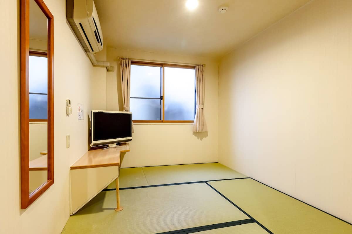A room with tatami flooring, a television on a wooden stand, a window with curtains, and a ceiling-mounted air conditioner.