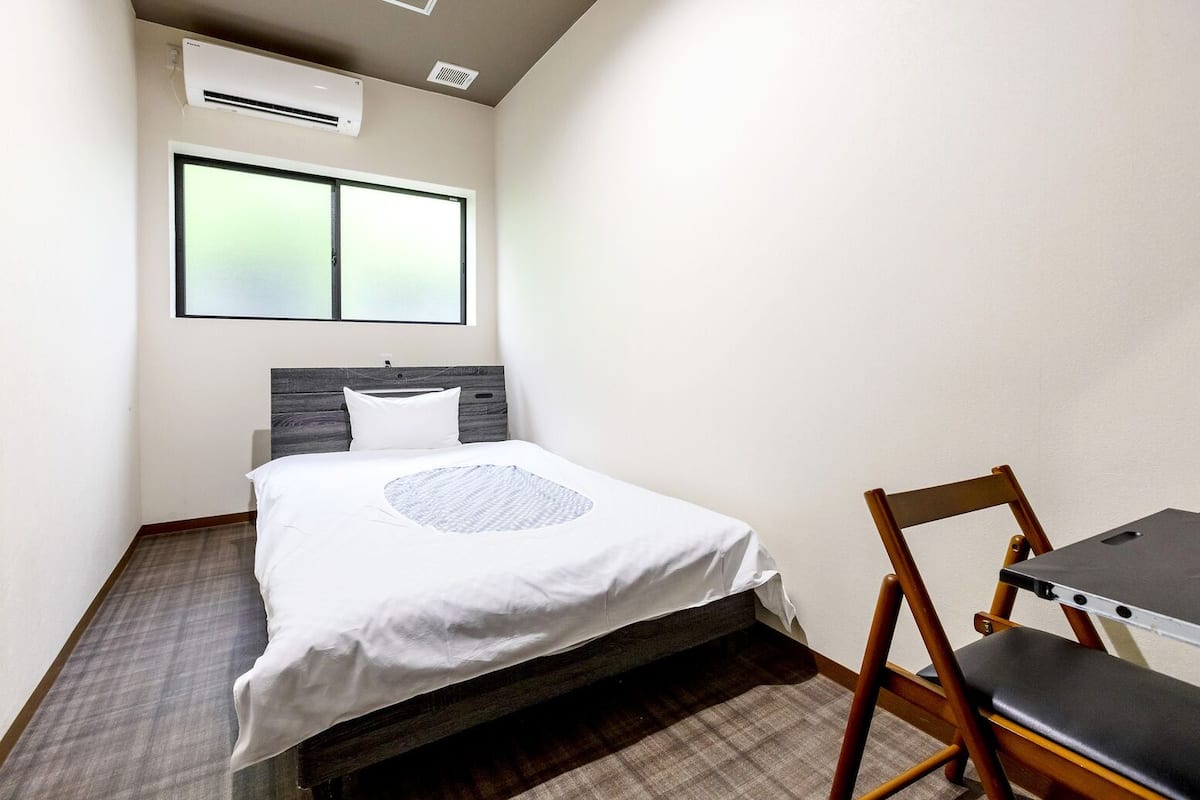 A small, neatly arranged room with a bed, a chair, a table, and a window.