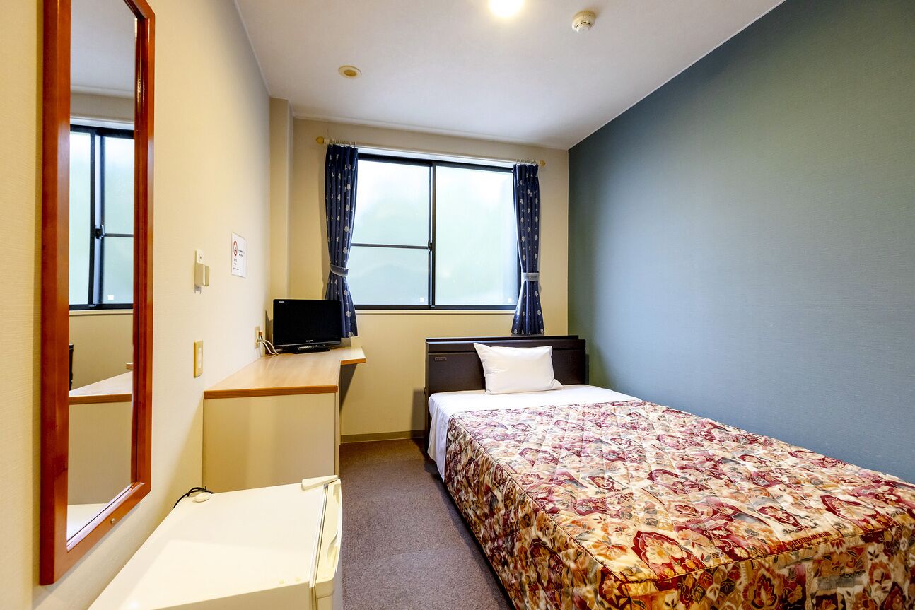 A hotel room with a bed, a desk with a computer, a window with curtains, and a mirror.
