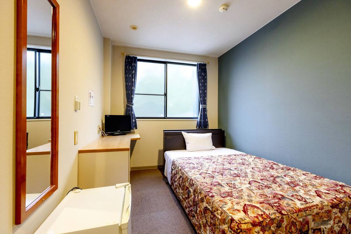 A hotel room with a bed, a desk with a computer, a window with curtains, and a mirror.
