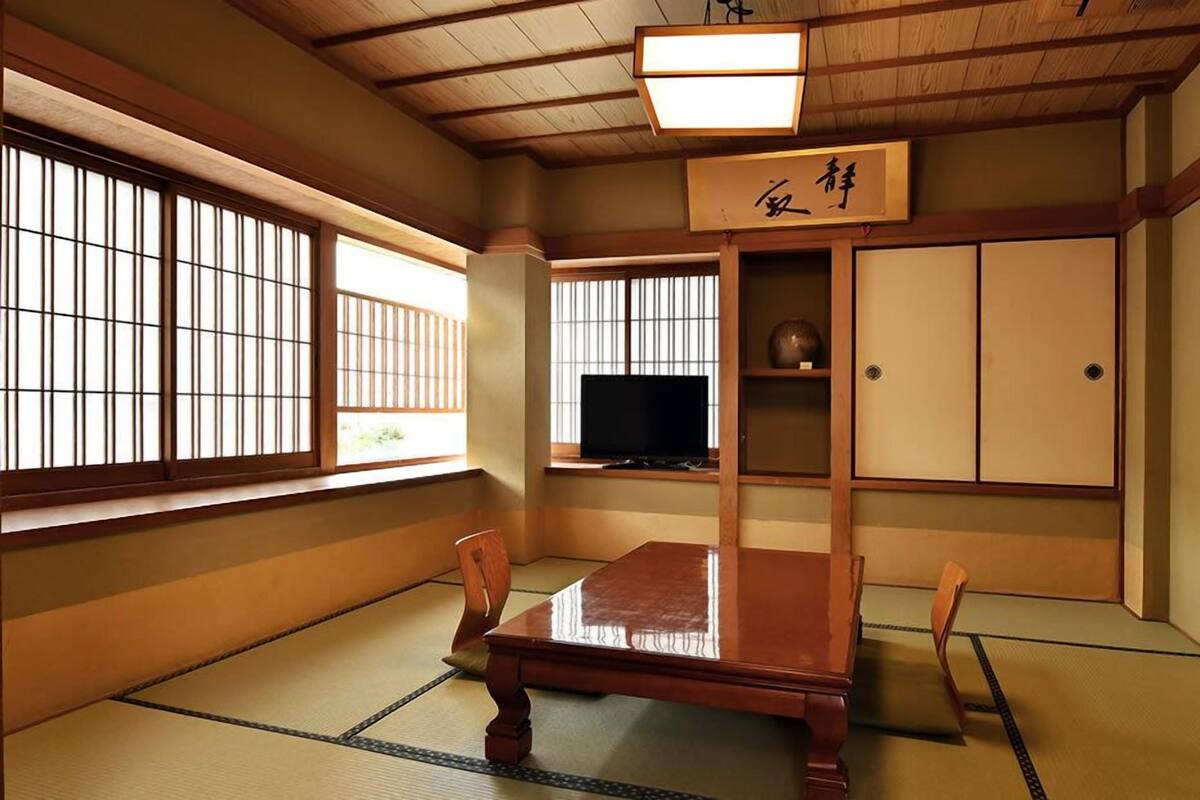 Ein traditioneller japanischer Raum mit Tatami-Boden, einem niedrigen Holztisch, einem Fernseher, Schiebetüren und einem Schild mit japanischen Schriftzeichen.