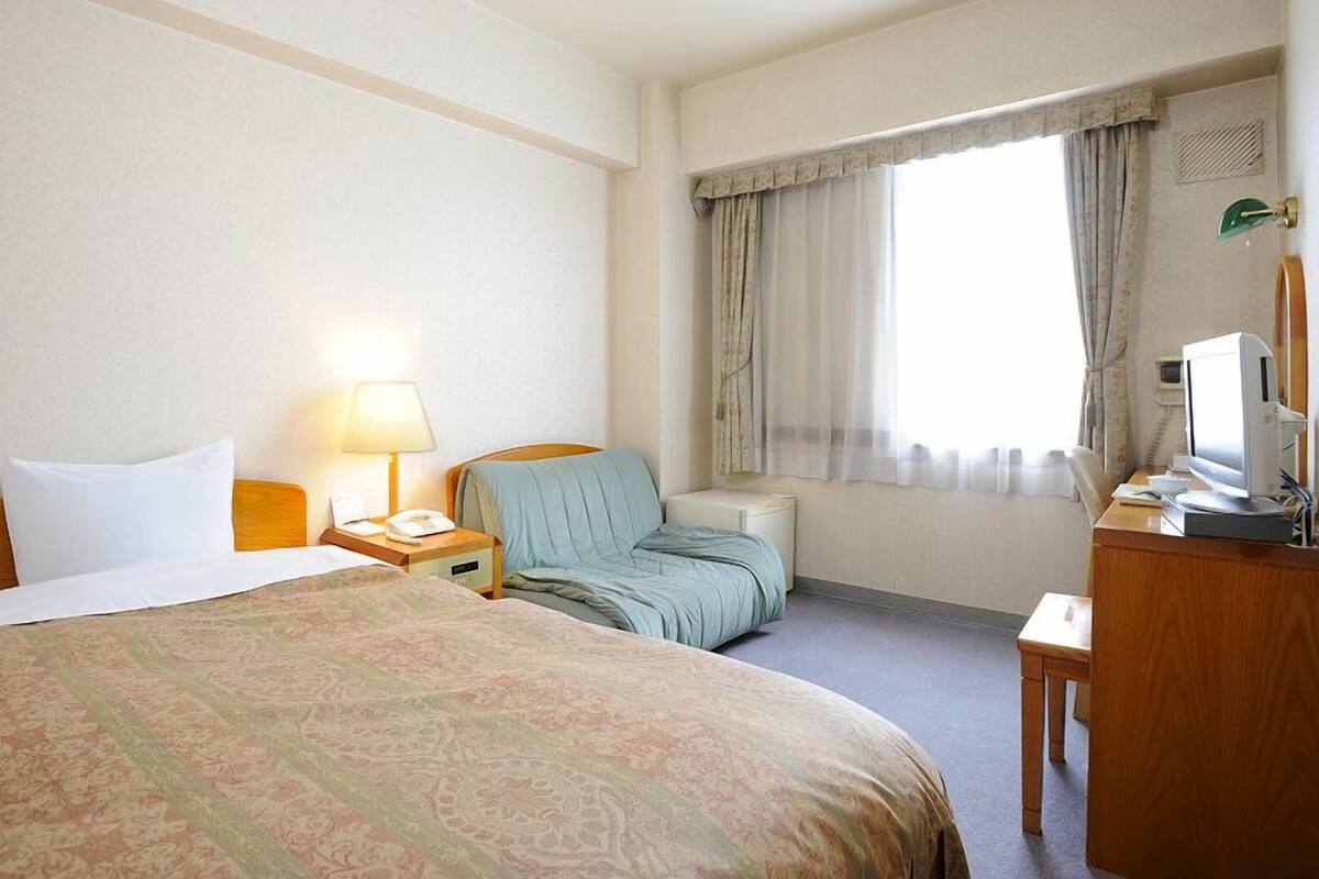 Une chambre d’hôtel avec un lit, un bureau avec un ordinateur, une chaise, une lampe et une fenêtre avec des rideaux.