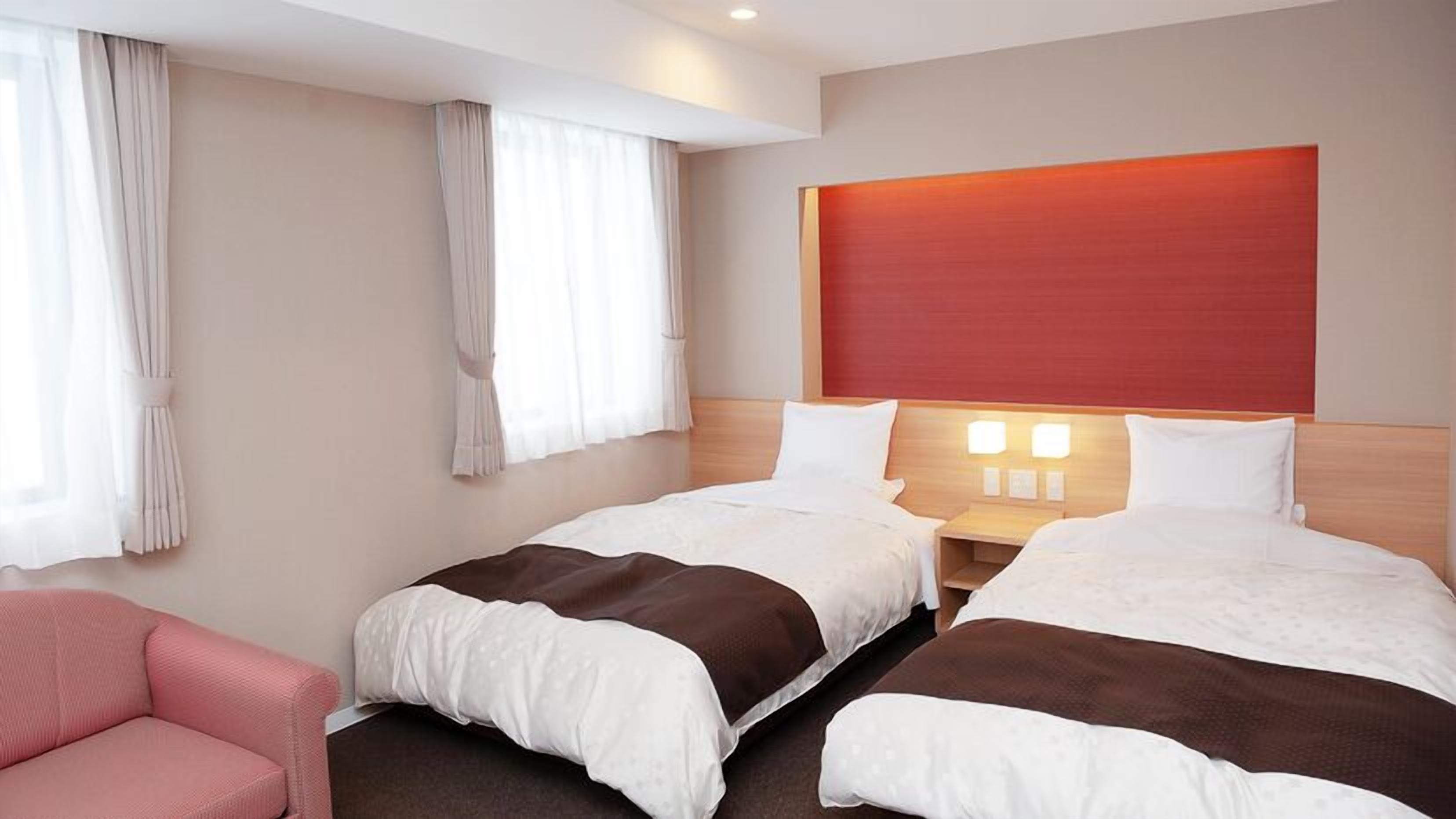 A hotel room with two beds, a wooden headboard, a nightstand with lamps, a pink chair, and sheer curtains.