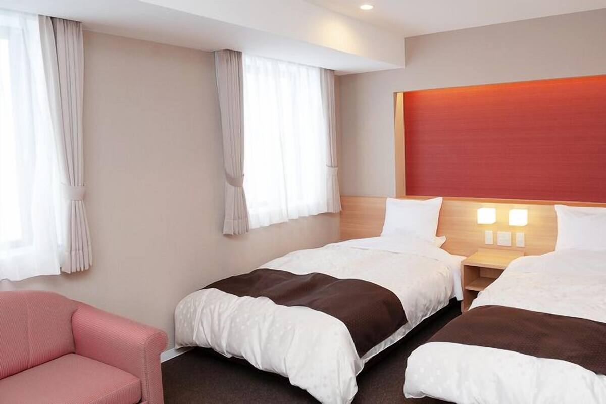 Une chambre d’hôtel avec deux lits, une tête de lit en bois, une table de chevet avec des lampes, une chaise rose et des rideaux légers.