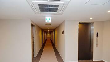 A hotel corridor with a central elevator, carpeted floor, and a ceiling vent.