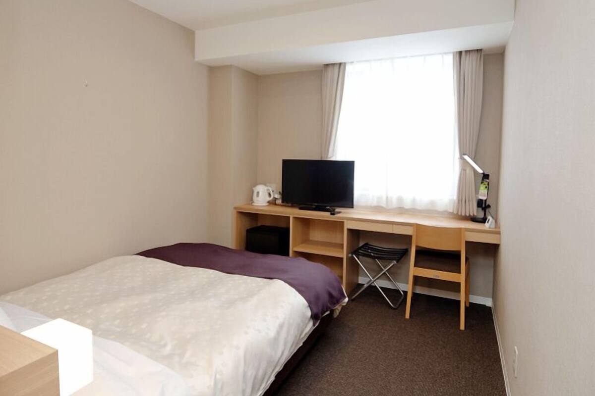 Une chambre d’hôtel avec un lit, un bureau avec une télévision, une chaise et une fenêtre avec des rideaux.