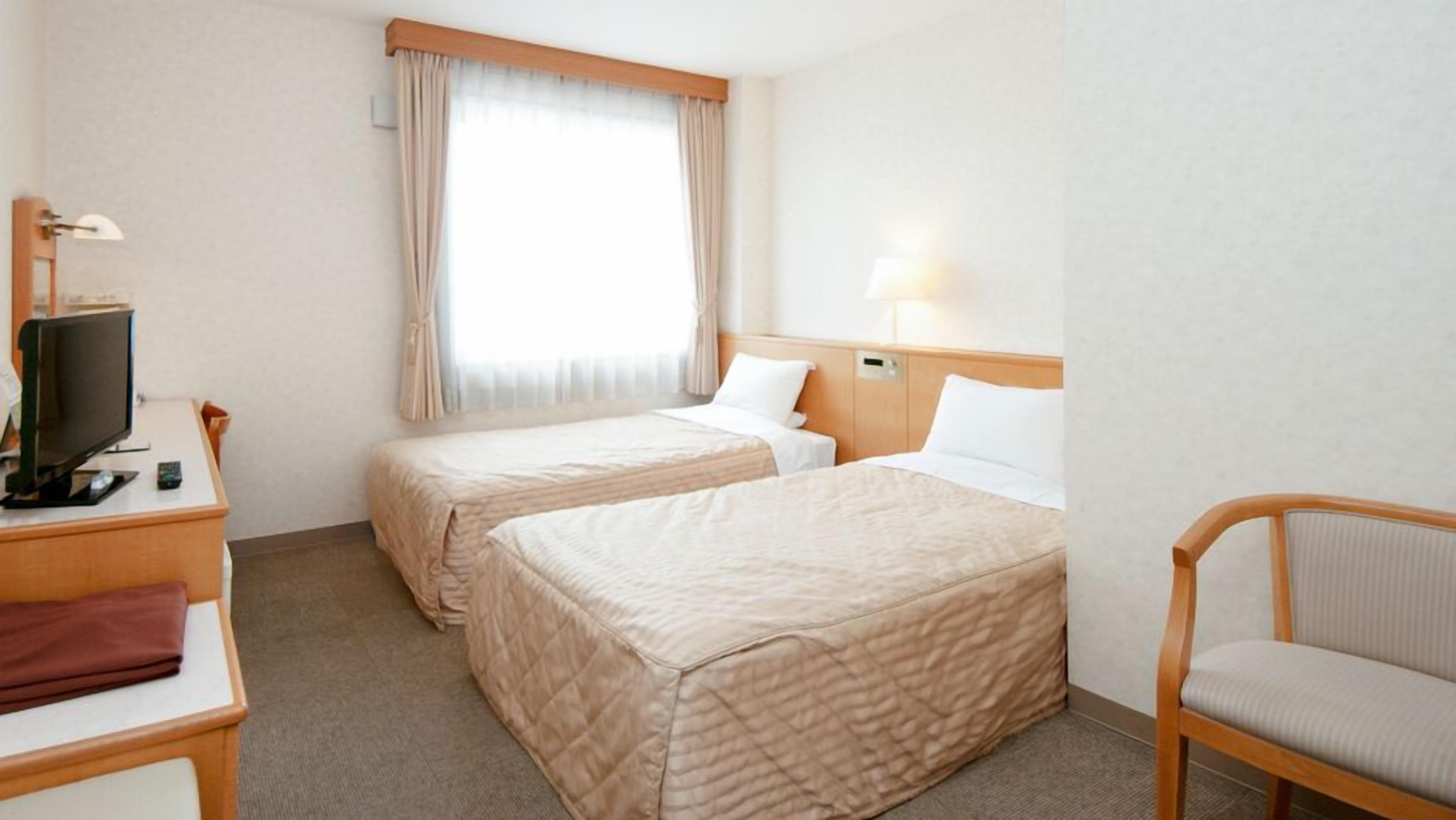A hotel room with two beds, a desk with a television, a chair, and a window with curtains.