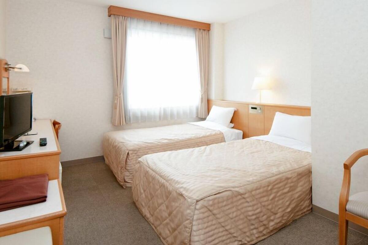 Une chambre d’hôtel avec deux lits, un bureau avec une télévision, une chaise et une fenêtre avec des rideaux.