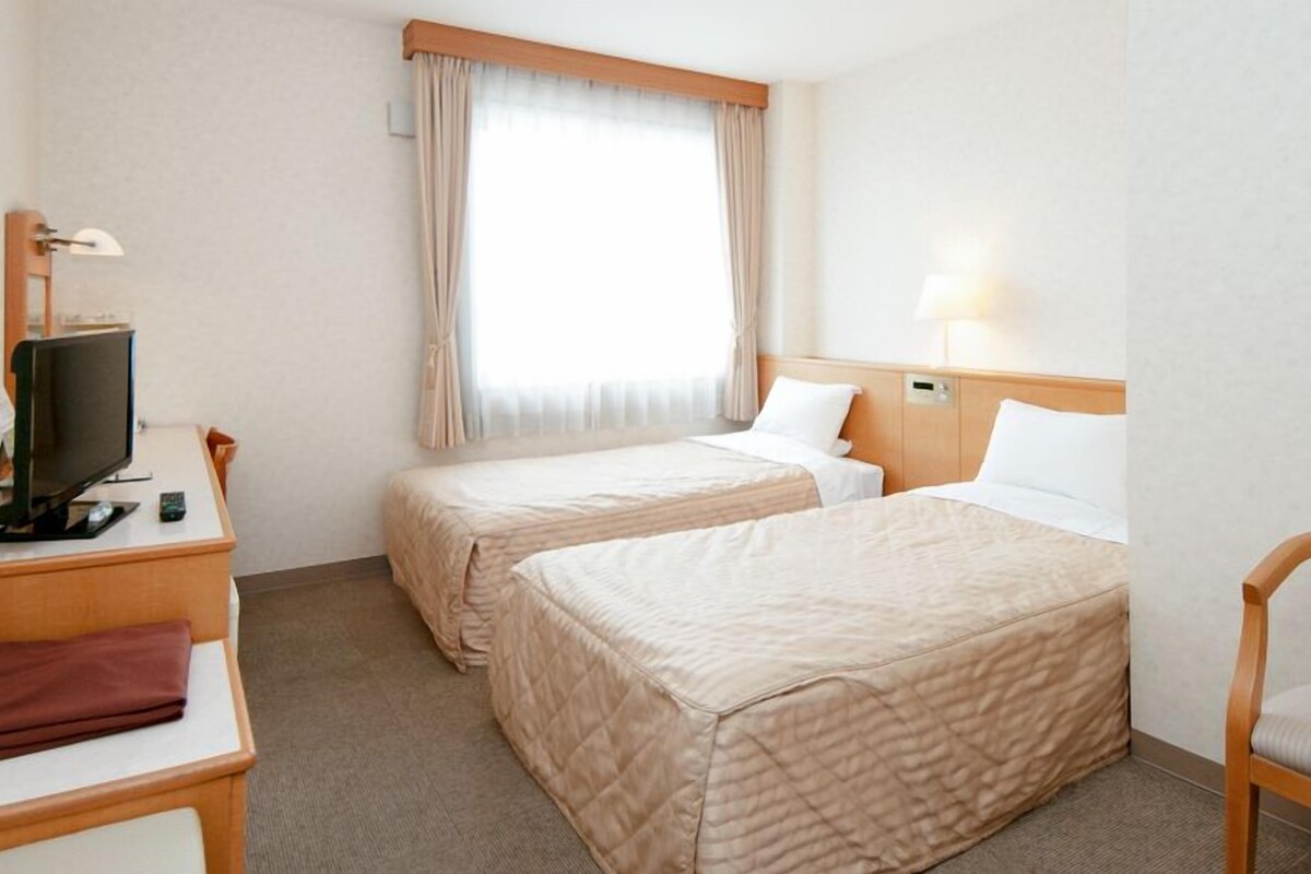 A hotel room with two beds, a desk with a television, a chair, and a window with curtains.