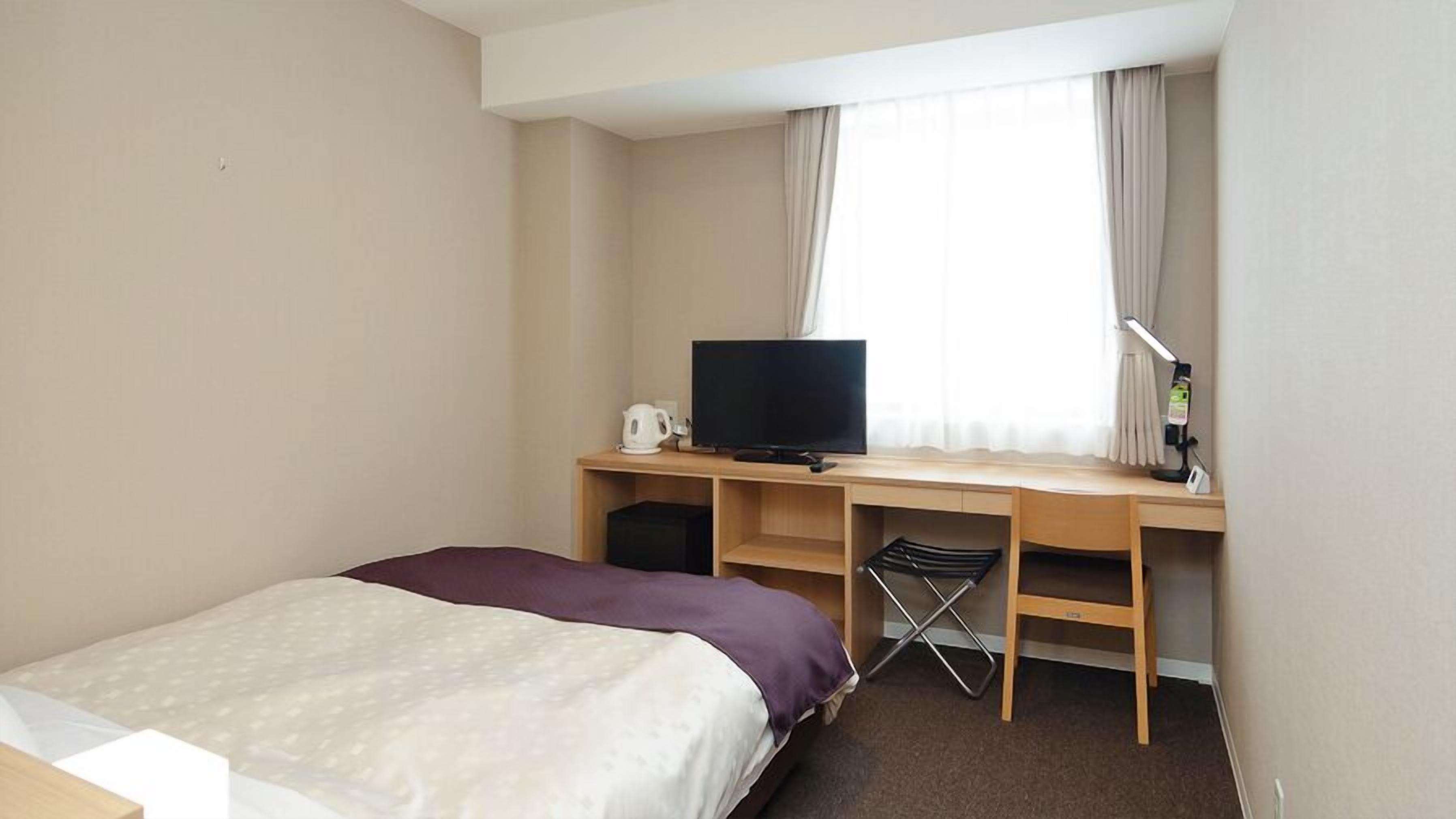 A hotel room with a bed, a desk with a television, a chair, and a window with curtains.