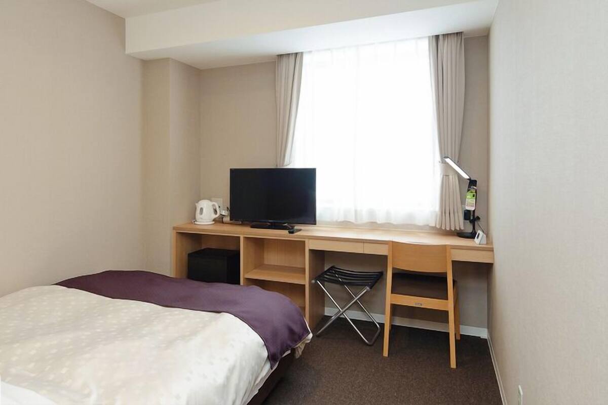 Une chambre d’hôtel avec un lit, un bureau avec une télévision, une chaise et une fenêtre avec des rideaux.