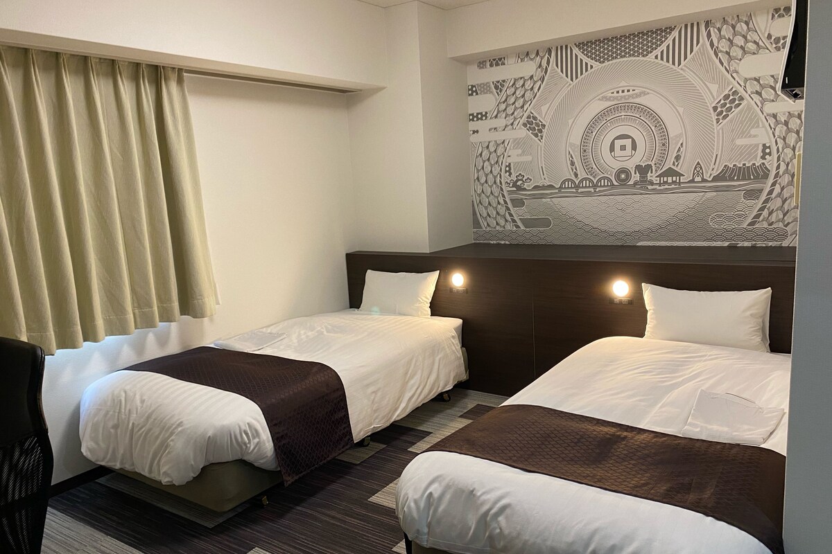 Ein Hotelzimmer mit zwei Betten, einem Kopfteil mit integrierter Beleuchtung, einem großen Wandteppich und einem Fenster mit Vorhängen.