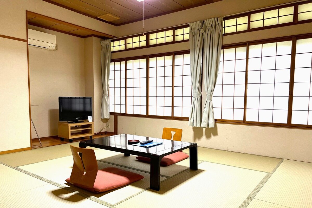 Una habitación tradicional japonesa con suelo de tatami, una mesa baja con cojines, un televisor sobre un soporte de madera y ventanas grandes con cortinas.