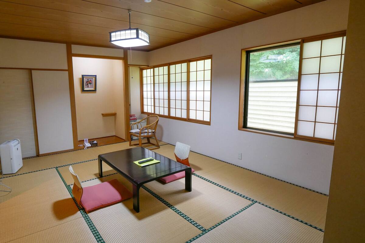 Una tradizionale stanza giapponese con pavimento in tatami, un tavolino con cuscini, una sedia, una finestra con paraventi shoji e una ventola a soffitto.