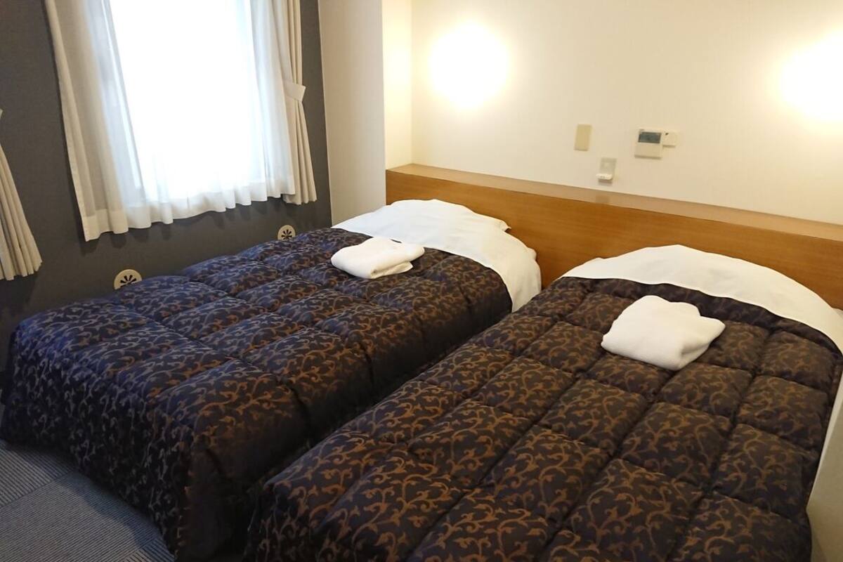 Two beds with patterned bedding, a wooden headboard, a wall-mounted light, and a window with curtains.