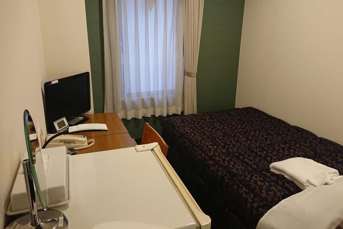 A hotel room with a bed, a desk with a computer, a telephone, and a window with curtains.