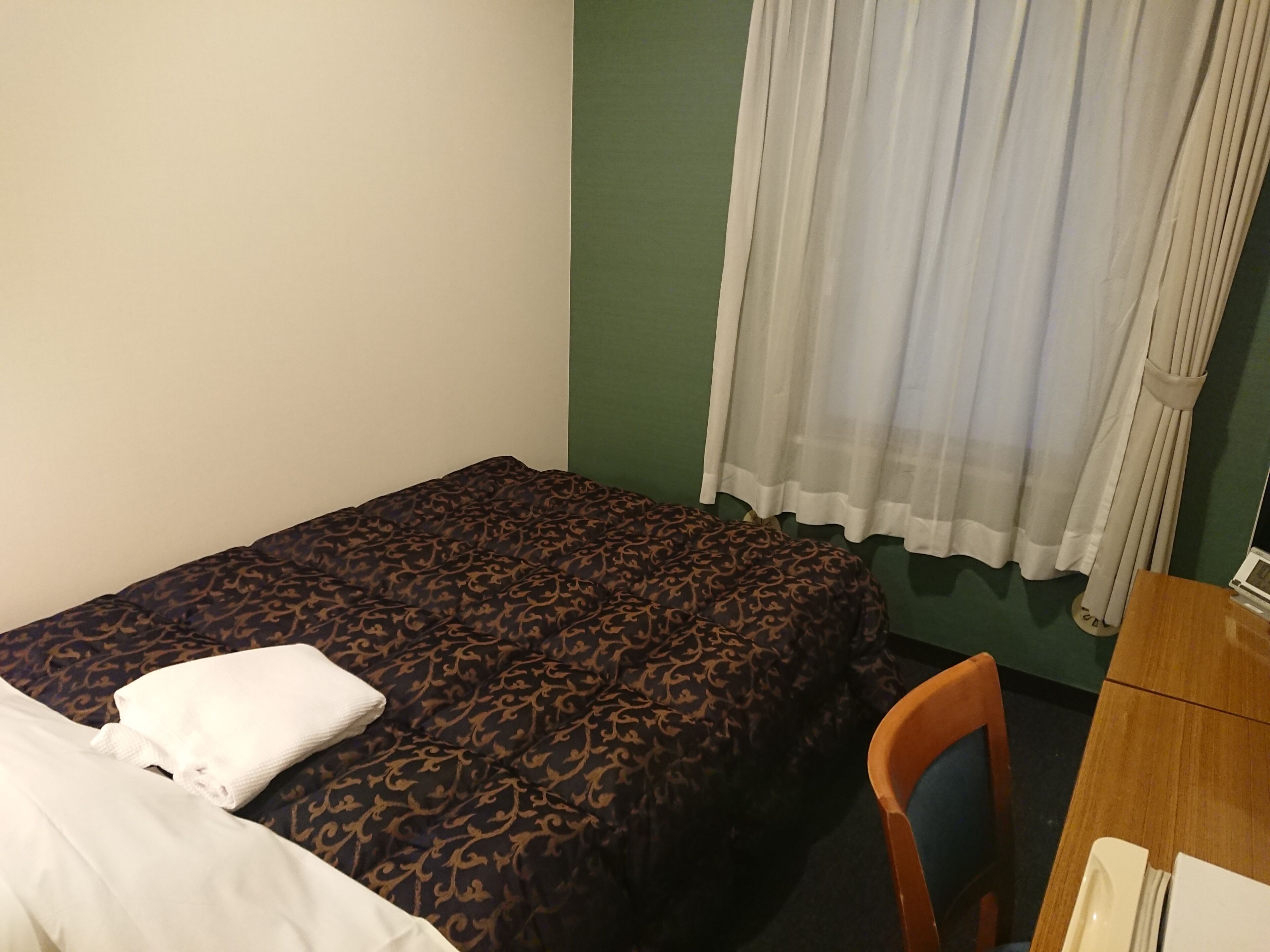 Una cama individual con una colcha estampada, una almohada blanca, una silla de madera y un escritorio con una lámpara.