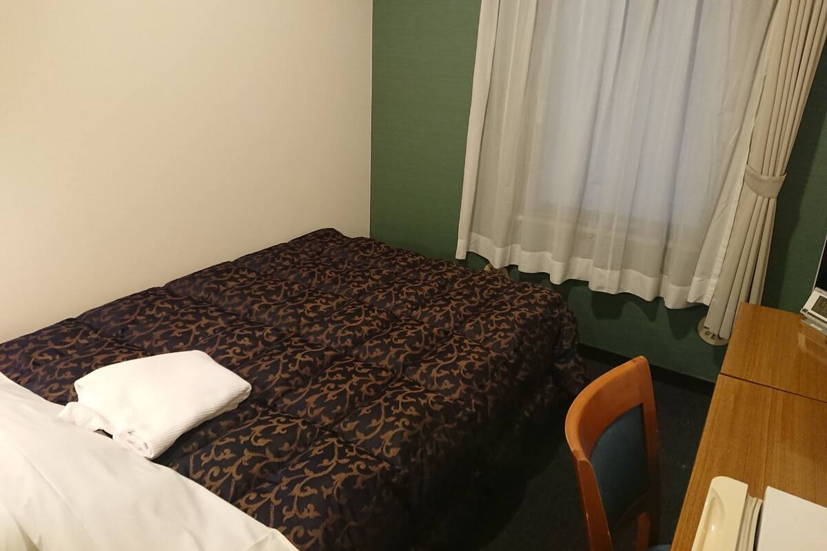 A single bed with a patterned bedspread, a white pillow, a wooden chair, and a desk with a lamp.