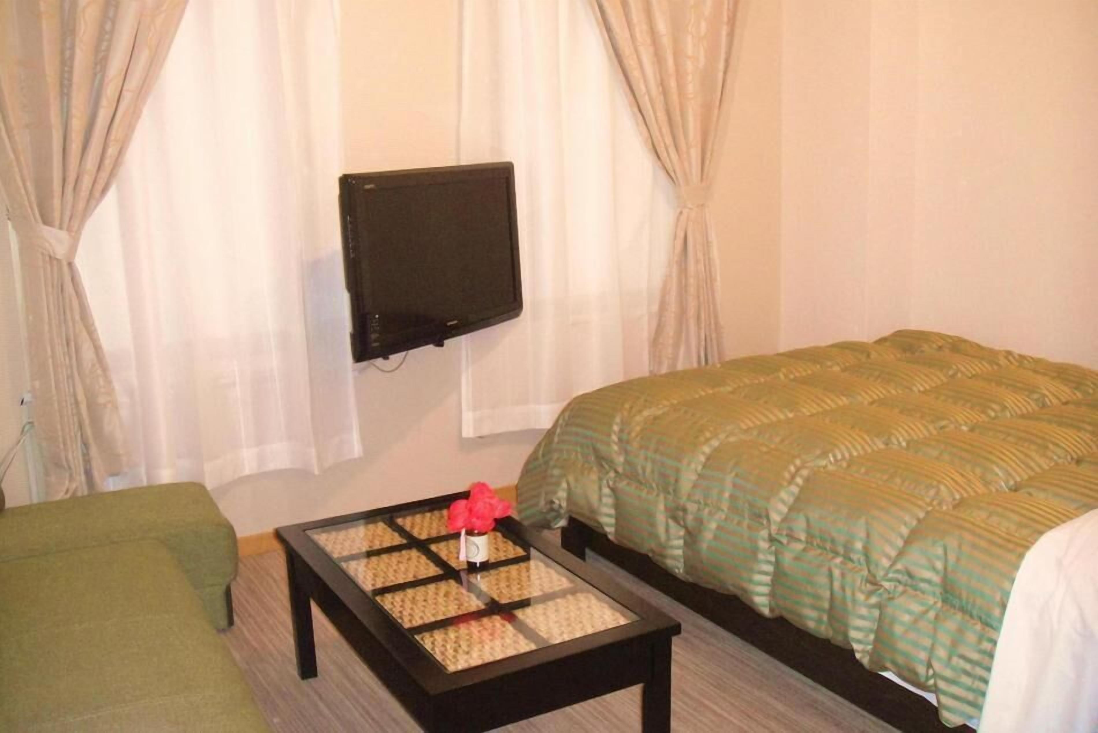 A hotel room with a bed, a green sofa, a small table with a vase, and a TV mounted on the wall.