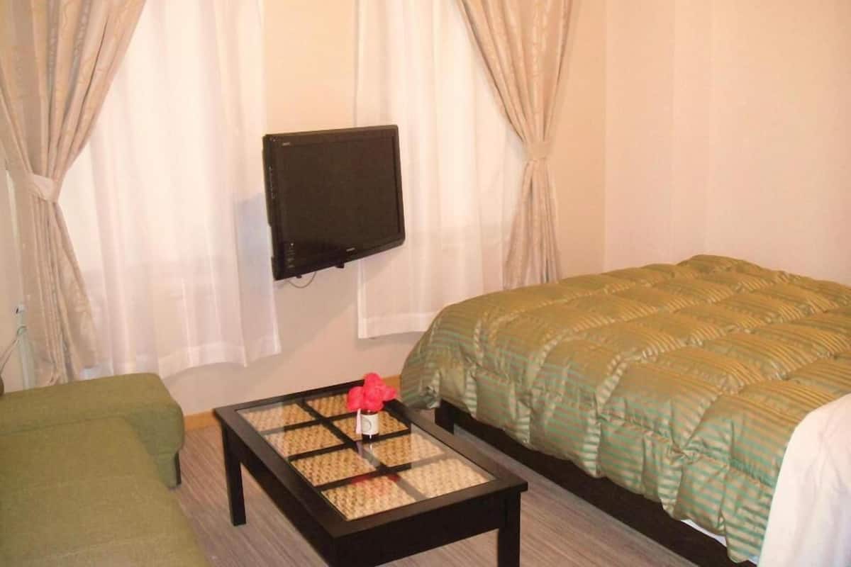 A hotel room with a bed, a green sofa, a small table with a vase, and a TV mounted on the wall.