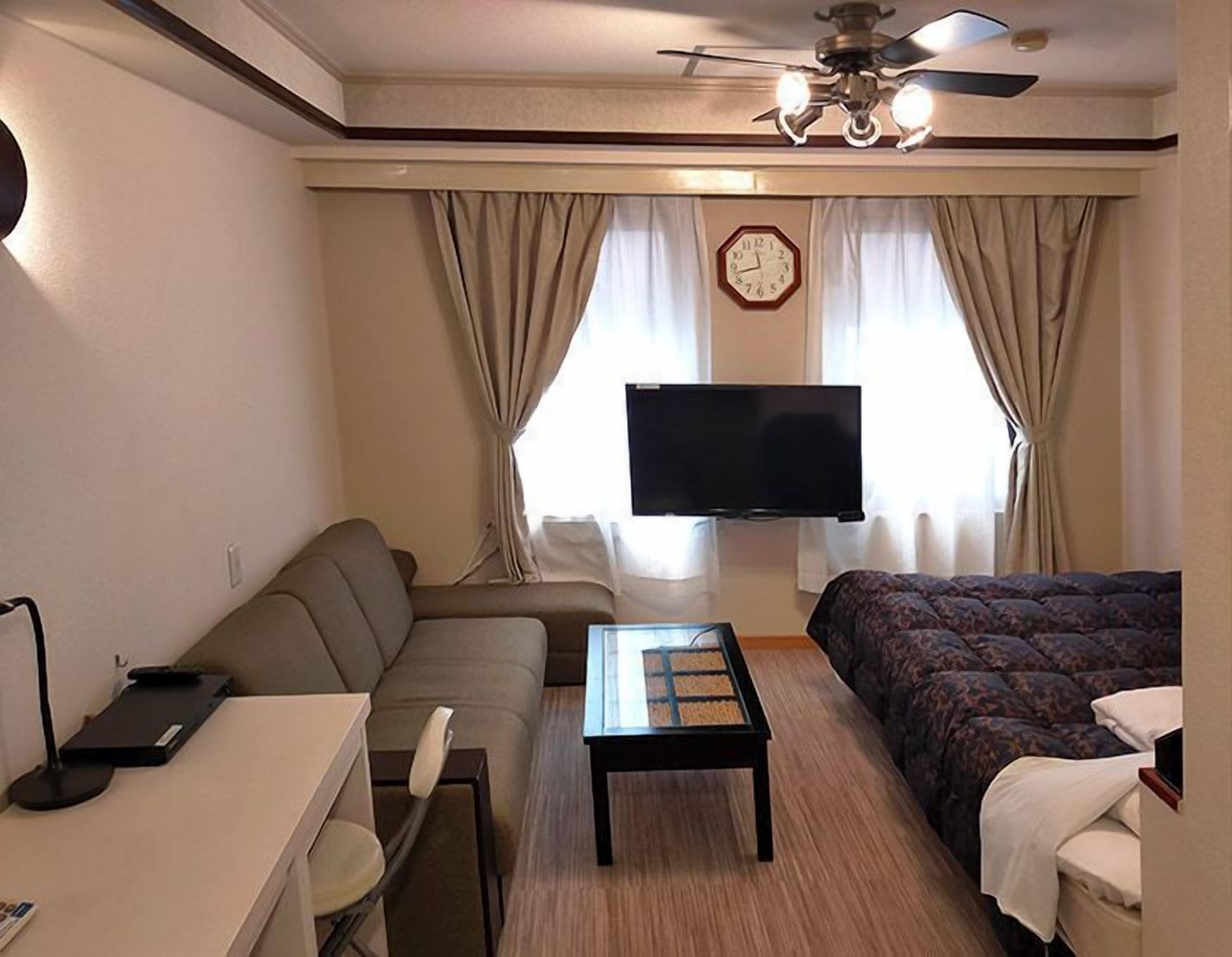 A hotel room with a sofa, a bed, a TV, a ceiling fan, and a clock on the wall.