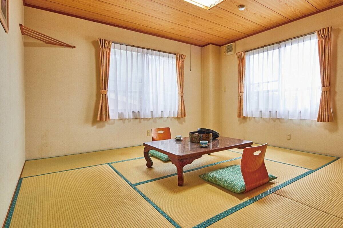 A traditional Japanese room with tatami flooring, a low wooden table, and two windows with curtains.