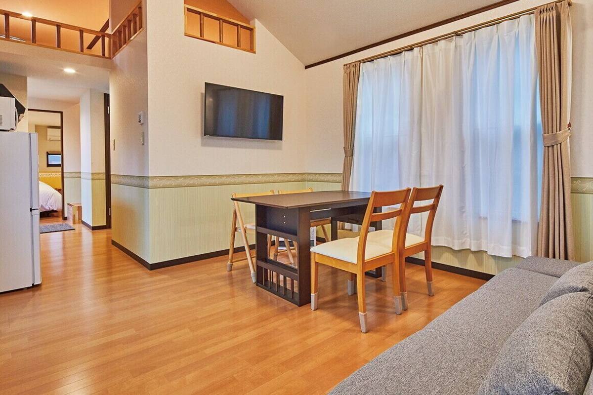 A dining area with a table and chairs, a television on the wall, and a window with curtains.