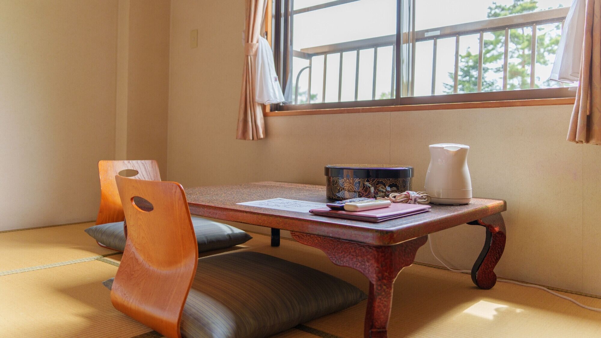 A traditional Japanese room with tatami flooring, a low wooden table, two chairs, a small table with a teapot, and a window with a view of greenery.