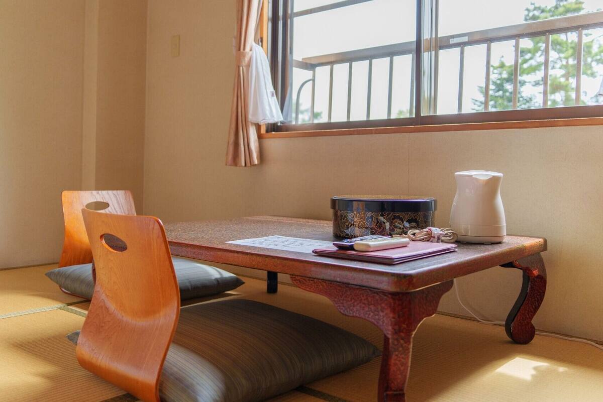 A traditional Japanese room with tatami flooring, a low wooden table, two chairs, a small table with a teapot, and a window with a view of greenery.