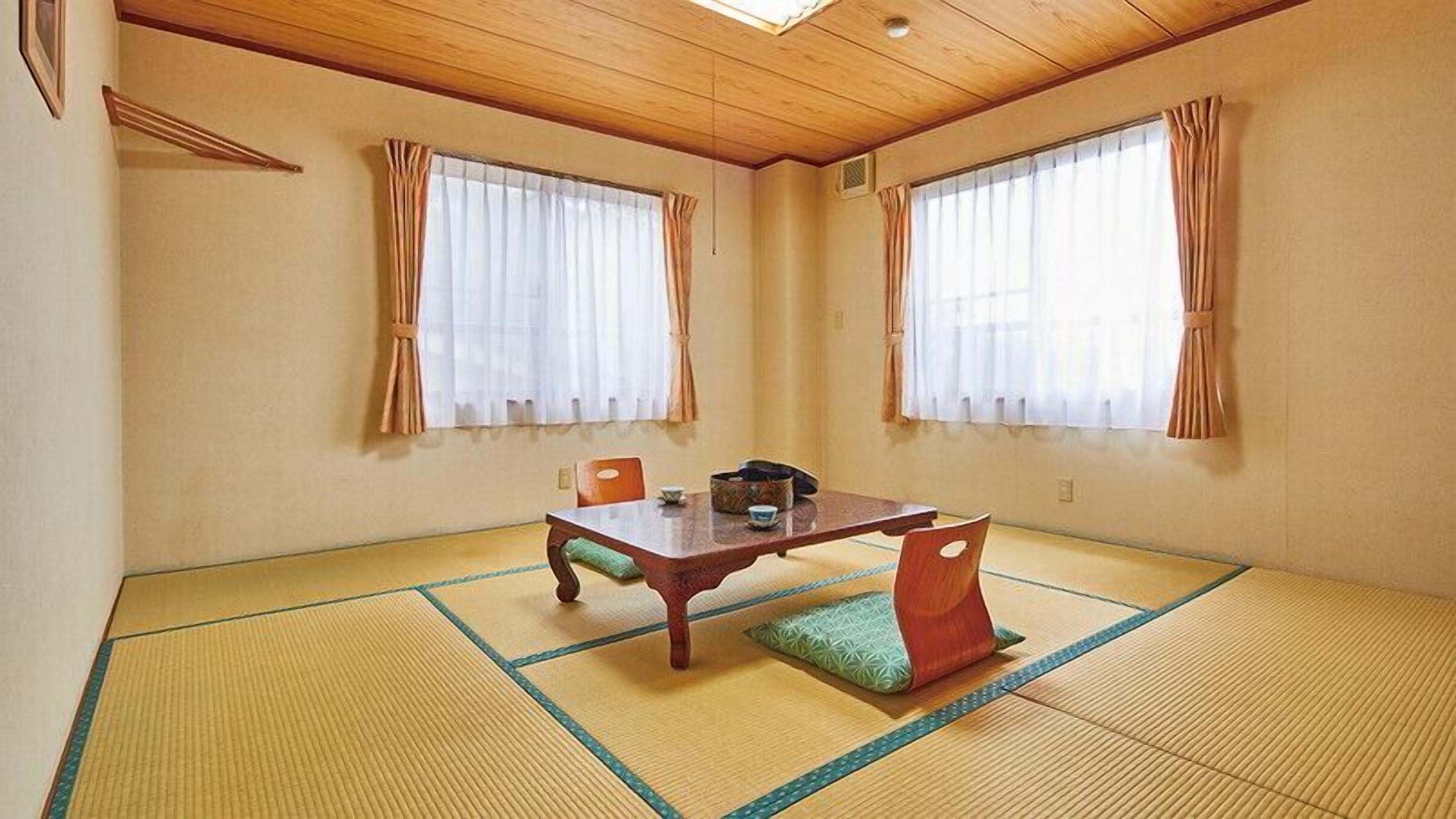 A traditional tatami mat room with a low table and chairs, large windows with curtains, and a wooden ceiling.