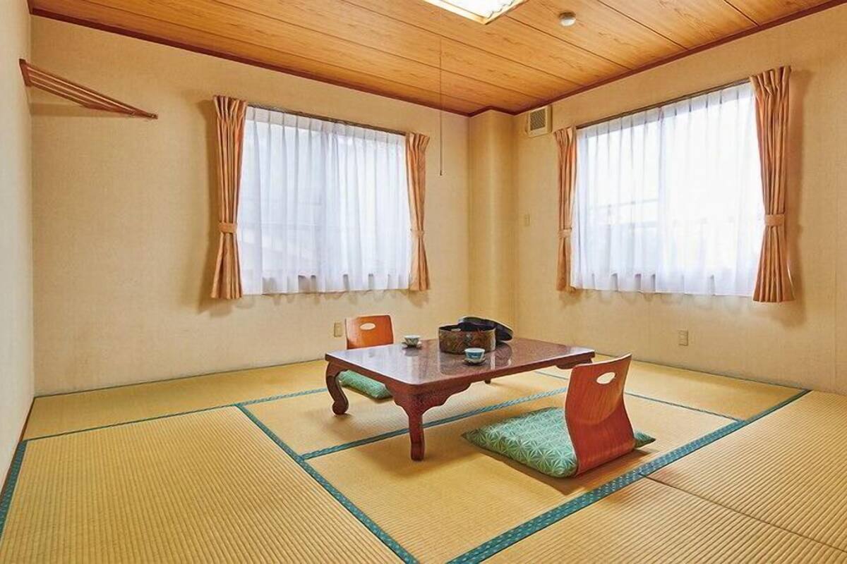 A traditional tatami mat room with a low table and chairs, large windows with curtains, and a wooden ceiling.