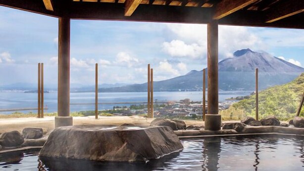 A hot spring area with a wooden structure overhead, a large rock in the center, and a view of a mountain and water body in the background. - Takarabaco Hot Spring Inn (Tarumizu)