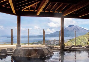 A hot spring area with a wooden structure overhead, a large rock in the center, and a view of a mountain and water body in the background. - Takarabaco Hot Spring Inn (Tarumizu)