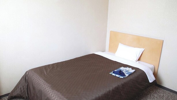 Een net opgemaakt bed met een bruine deken en een wit kussen in een eenvoudige kamer met effen muren.