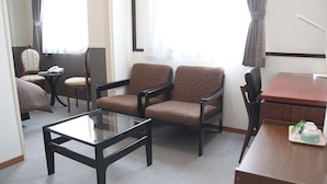 Hotelkamer met een tafel met glazen blad, twee bruine fauteuils, een houten bureau, een stoel en een klein tafeltje met een groen voorwerp erop.