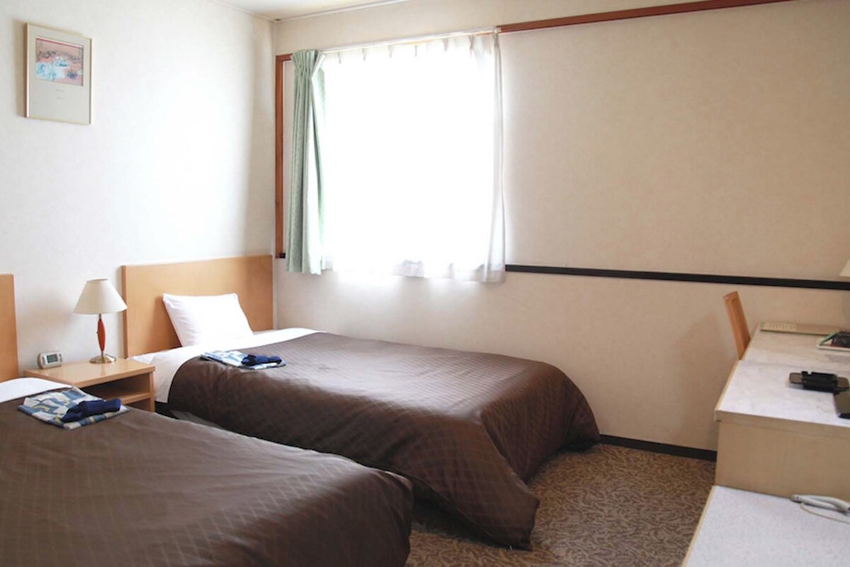 A hotel room with two beds, a desk, a chair, a nightstand, a lamp, a window with curtains, and a framed picture on the wall.