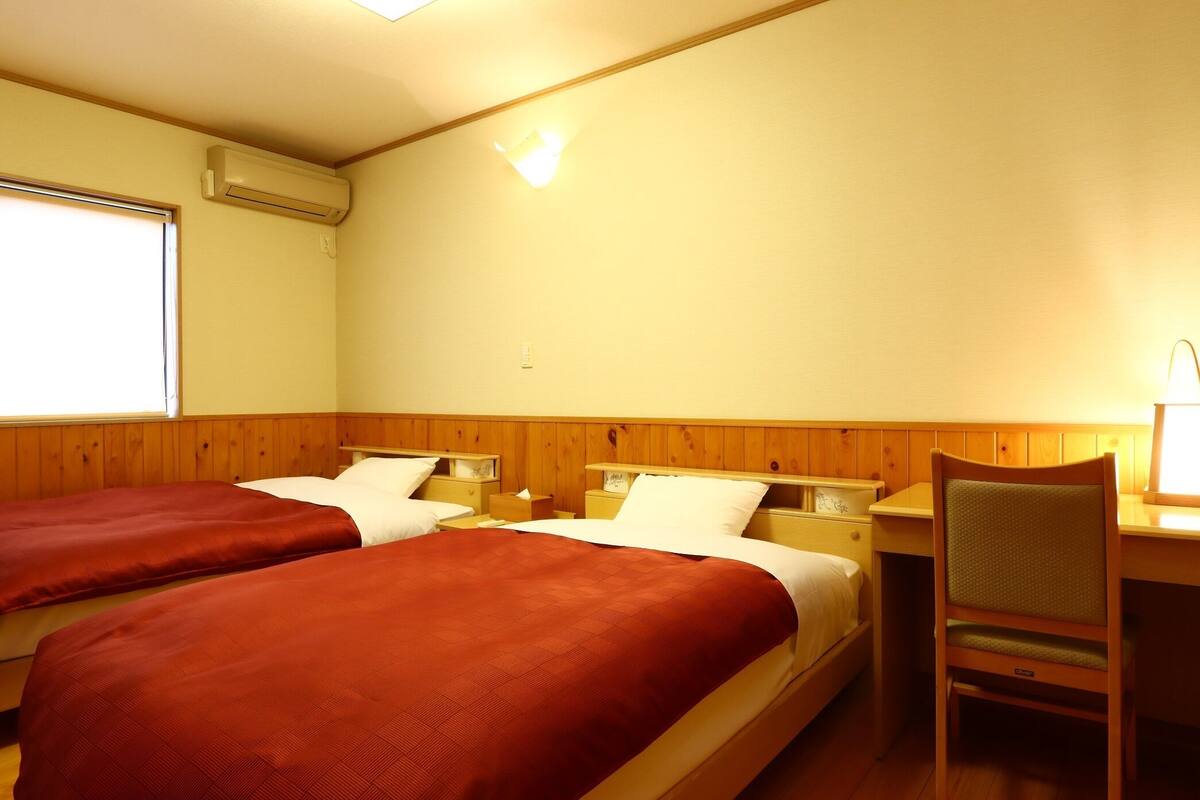 A hotel room with two beds, wooden paneling, a desk with a chair, and an air conditioning unit.