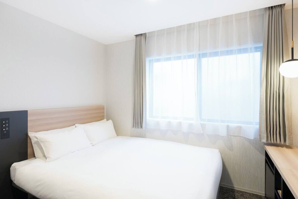 A hotel room with a bed, a control panel, a desk, and a window with curtains.