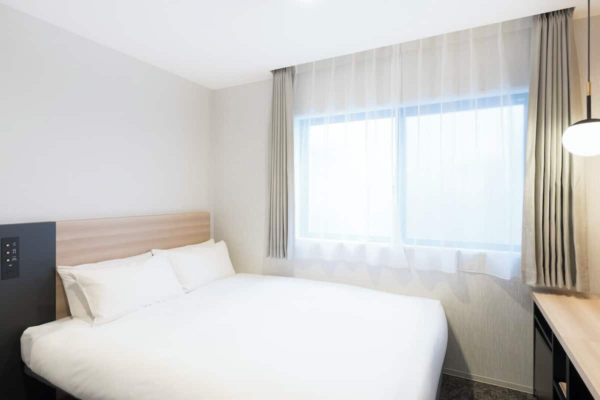 A hotel room with a bed, a control panel, a desk, and a window with curtains.