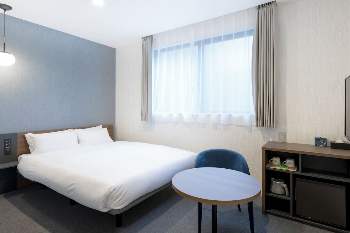 A hotel room with a bed, bedside table, chair, TV, and a window with curtains.