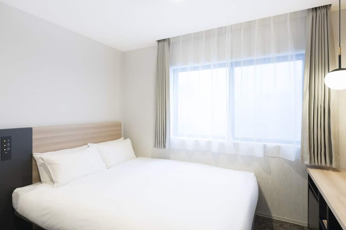 A hotel room with a bed, a control panel, a desk, and a window with curtains.