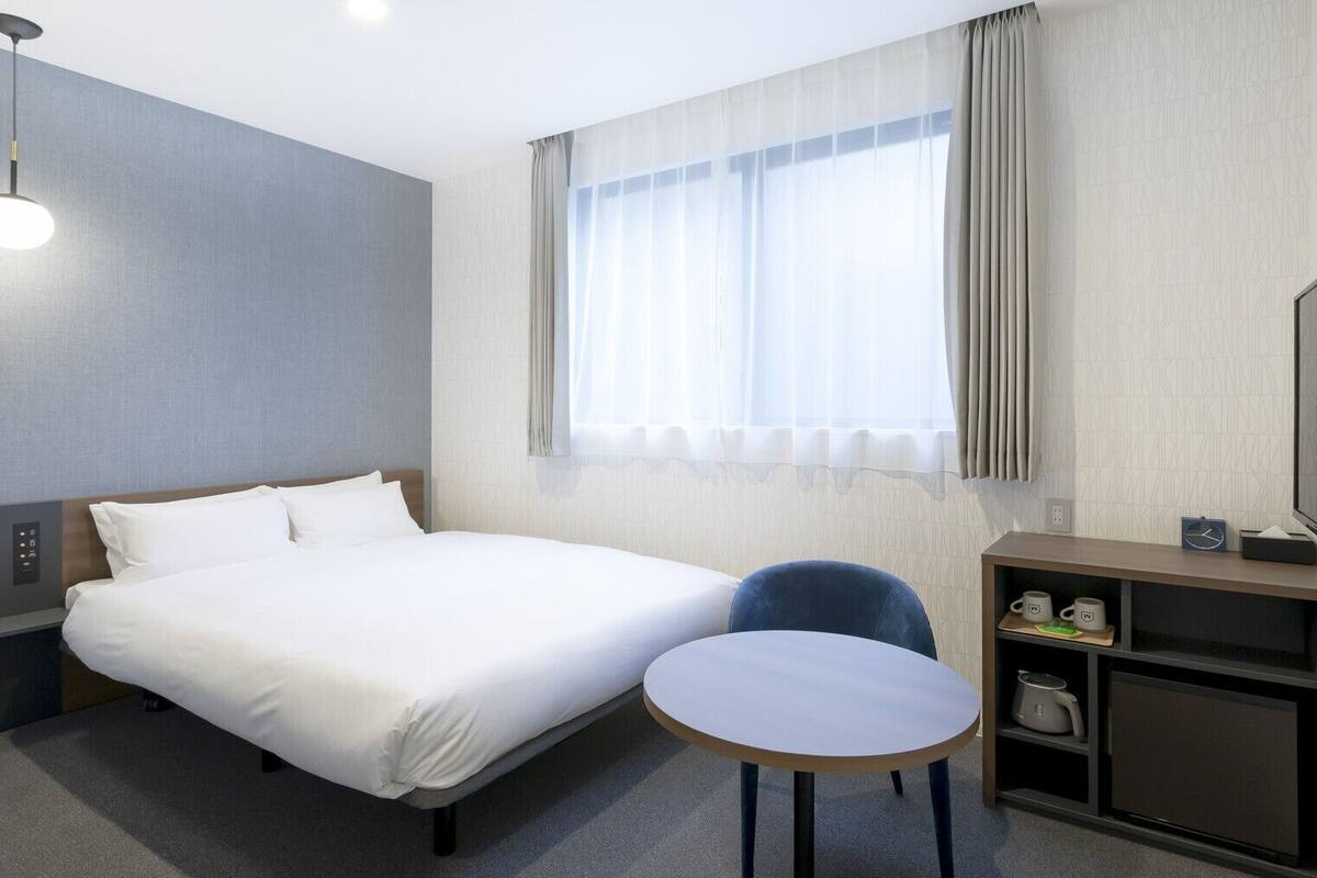 A hotel room with a bed, a chair, a small table, a TV, and a window with curtains.