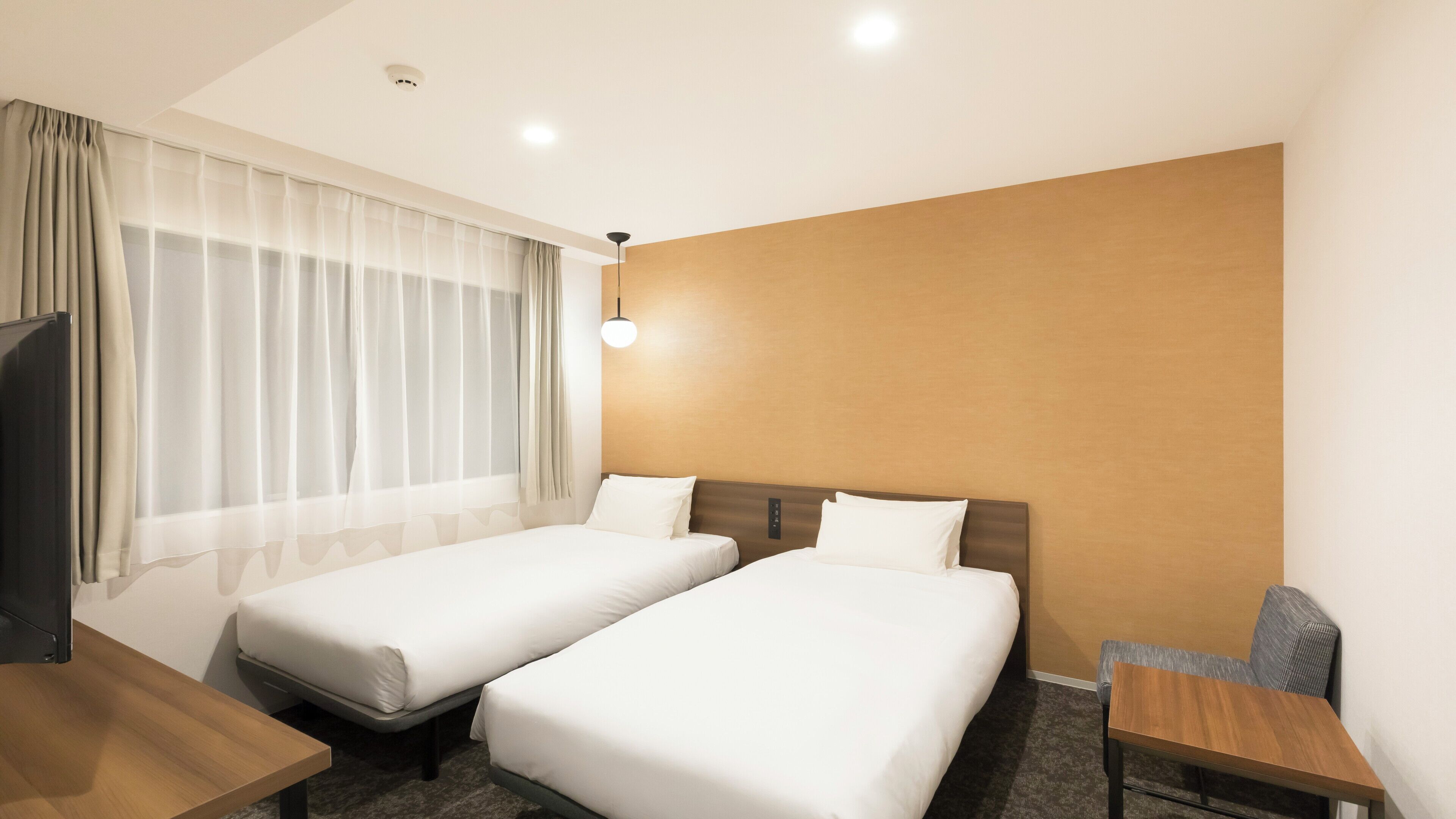 Ein Hotelzimmer mit zwei Einzelbetten, einem Schreibtisch, einem Stuhl, einem Fernseher und einem Fenster mit leichtem Vorhang.