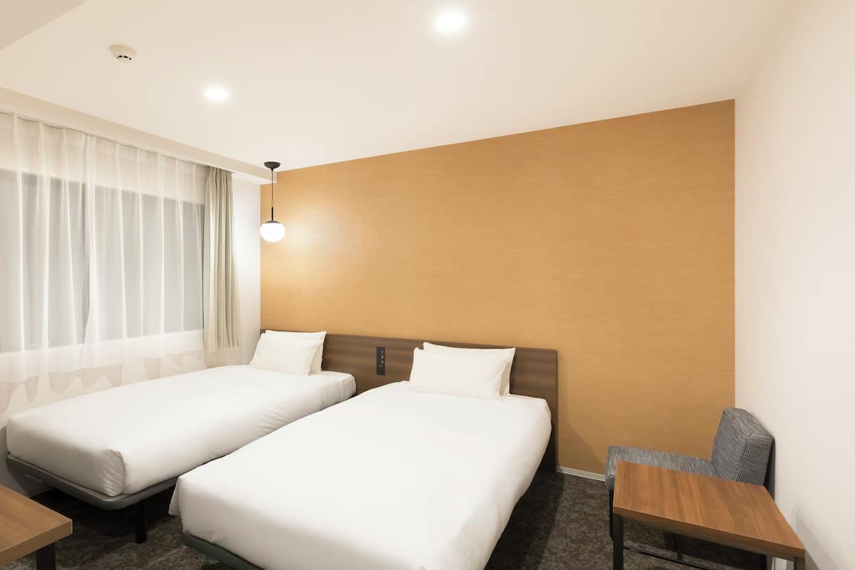 A hotel room with two single beds, a desk, a chair, a TV, and a window with sheer curtains.